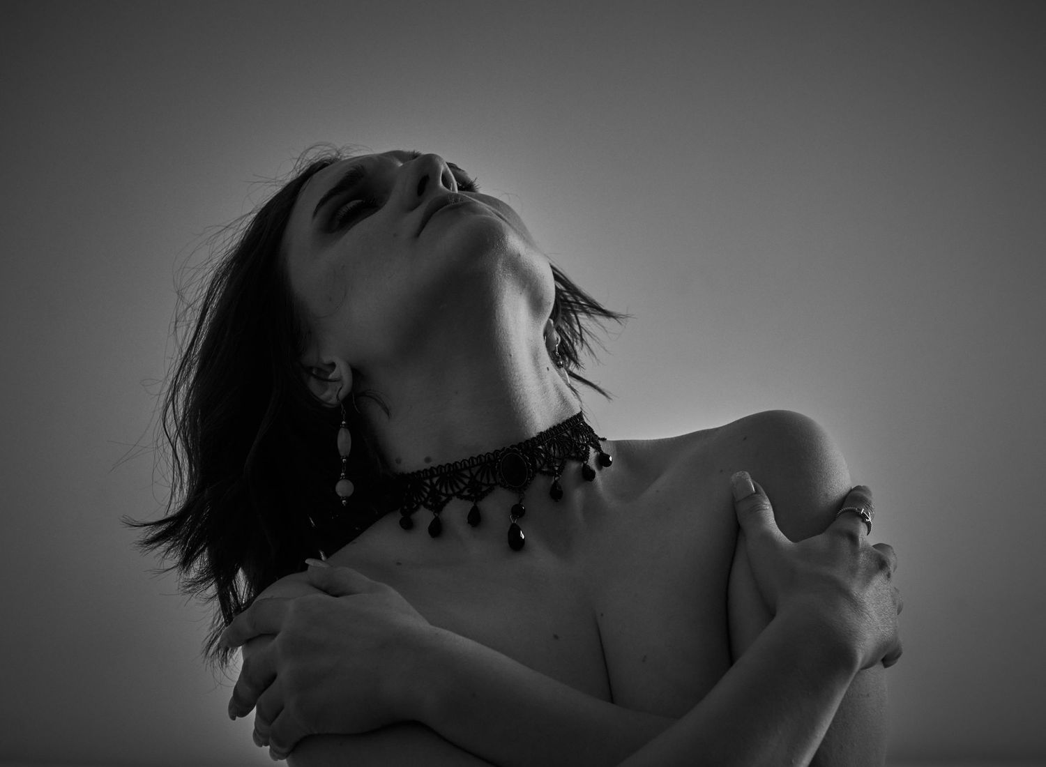 Black and white artistic portrait of woman with choker necklace hugging herself with head tilted back.