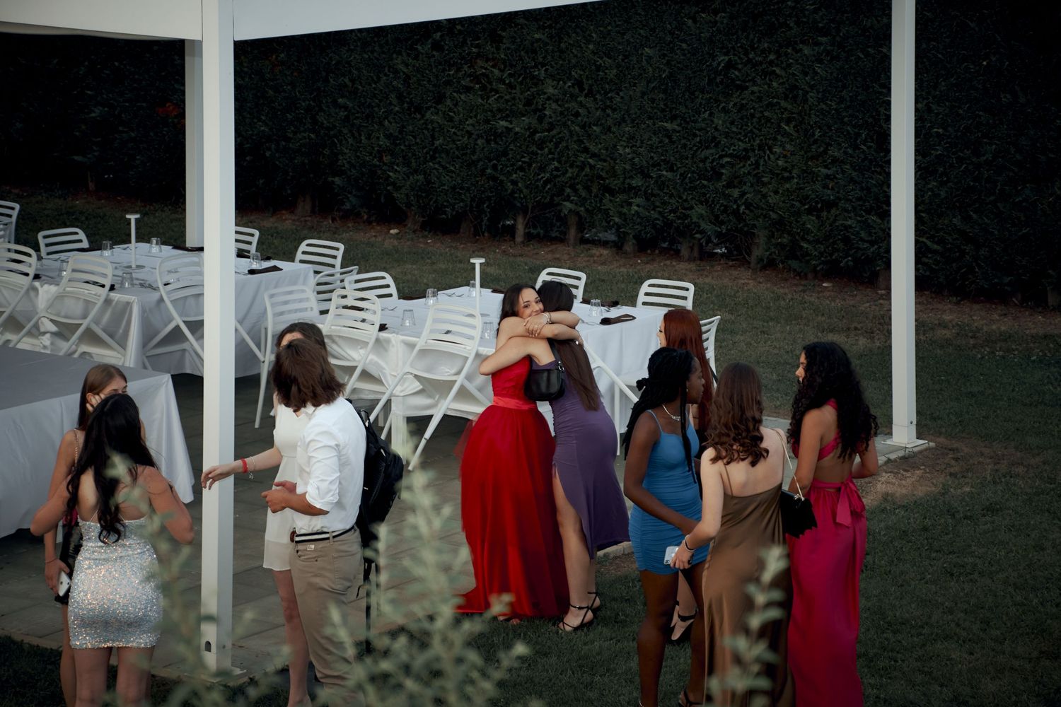 Group of elegantly dressed young women gather outdoors near white chairs at an evening event.