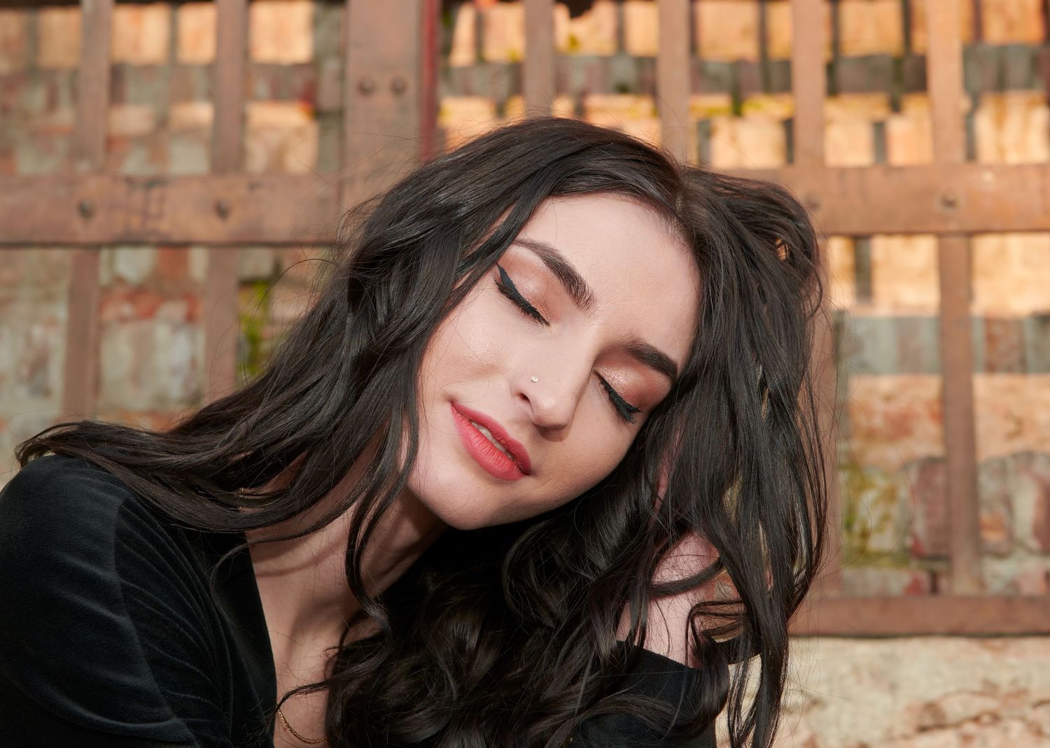 A beautiful young woman with dark wavy hair and red lips poses with eyes closed outdoors.