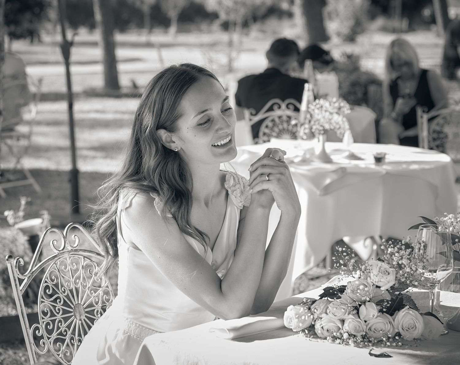 Smiling bride in white dress sitting at an outdoor wedding reception table with flowers.