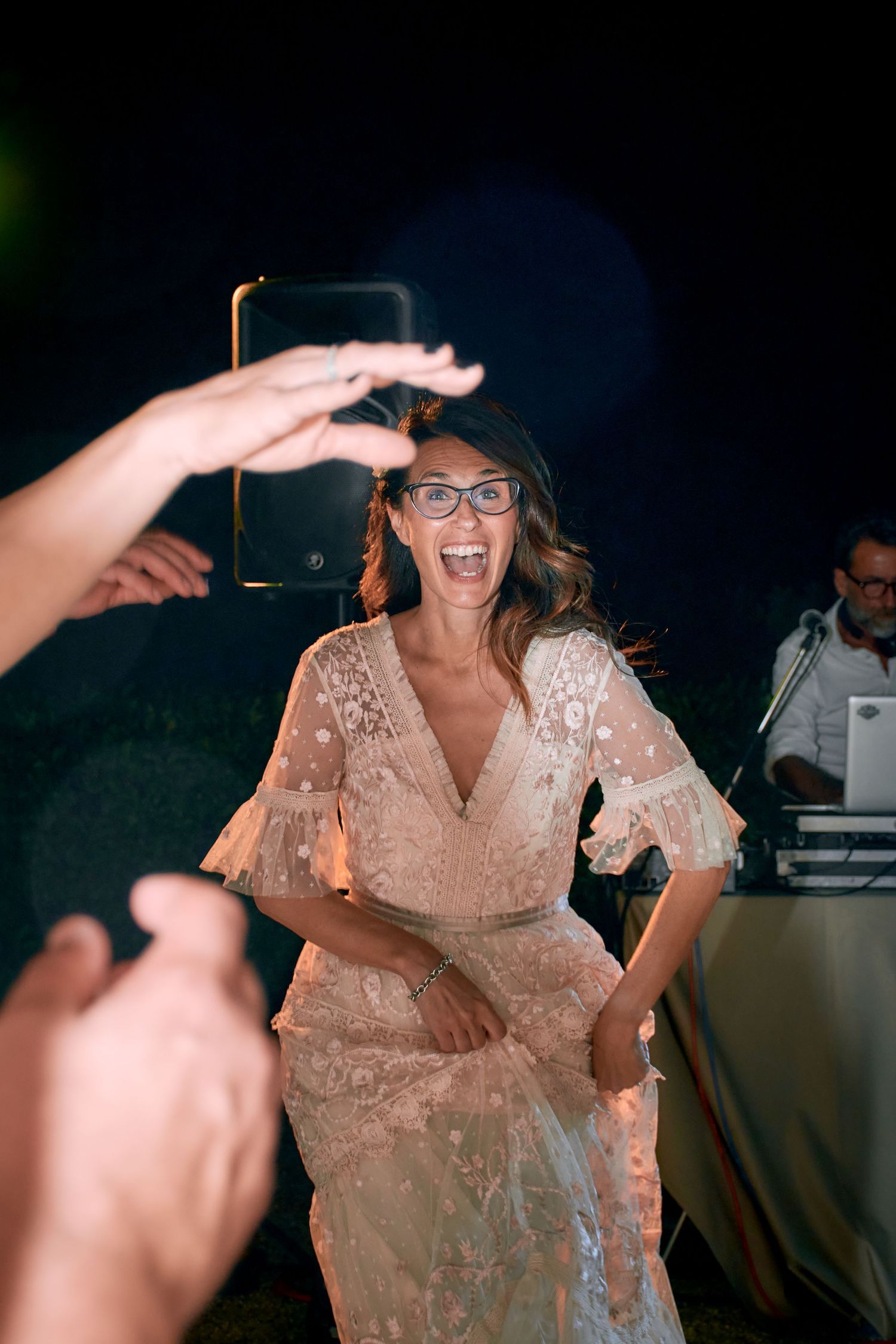 Excited bride in lace dress wearing glasses dancing joyfully at her evening wedding reception.