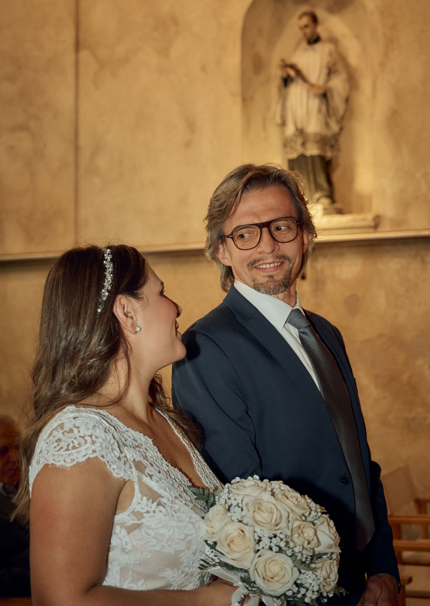 Bride holding white rose bouquet smiling at groom during church wedding ceremony.