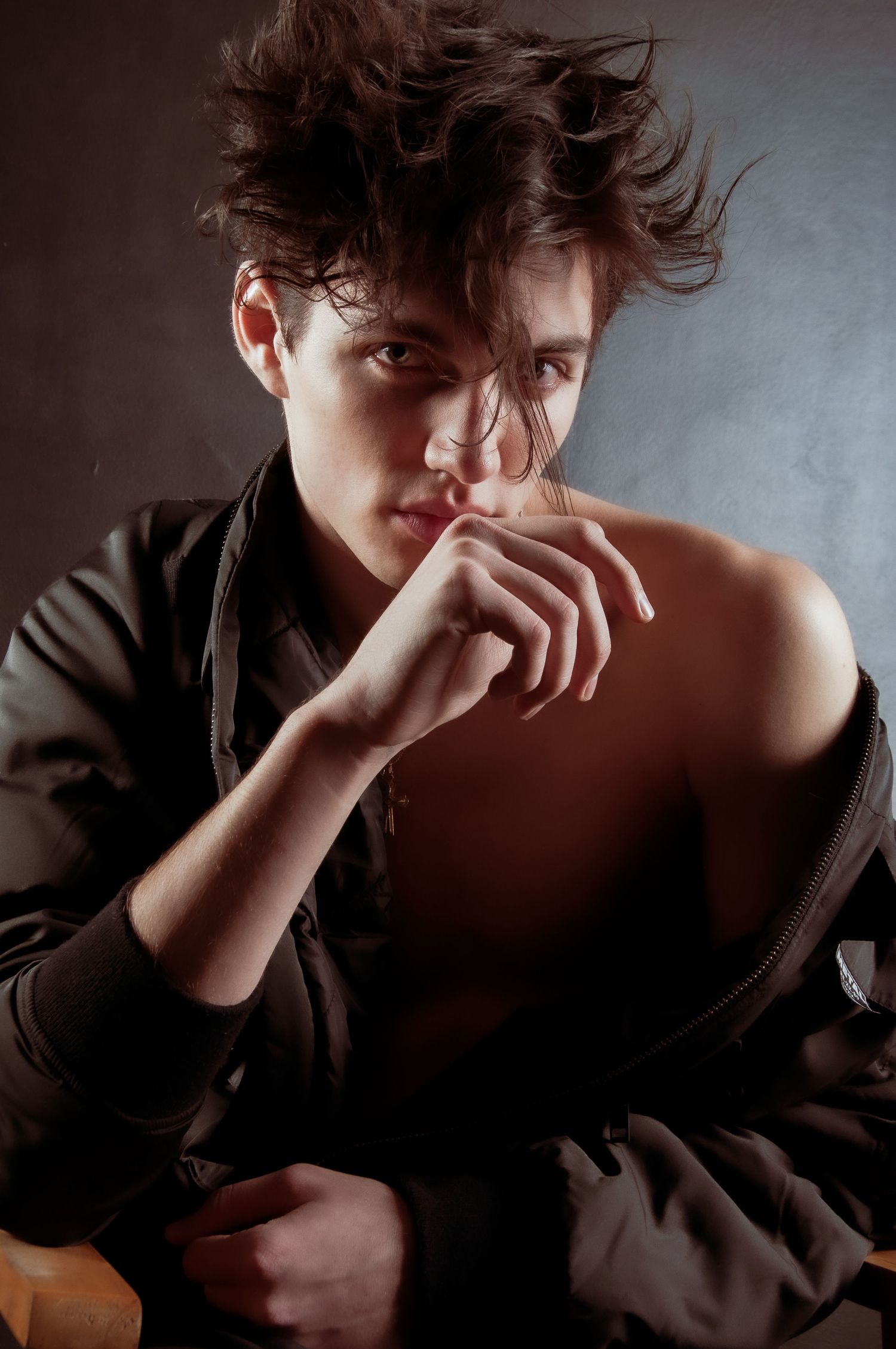Young male model with windswept dark hair wearing open jacket in dramatic studio lighting.