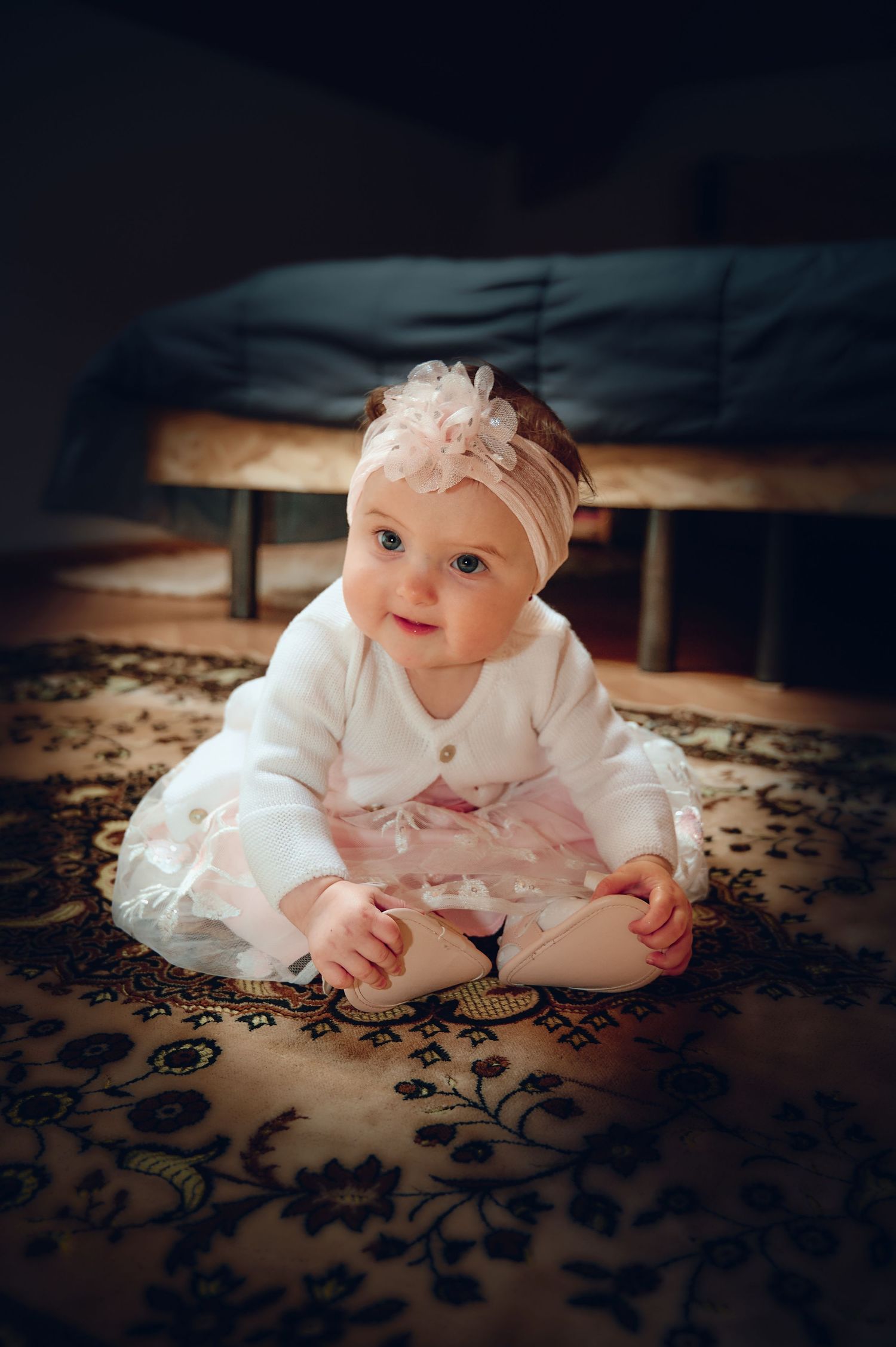 Adorable baby girl in white cardigan and pink tutu skirt sitting on patterned rug.