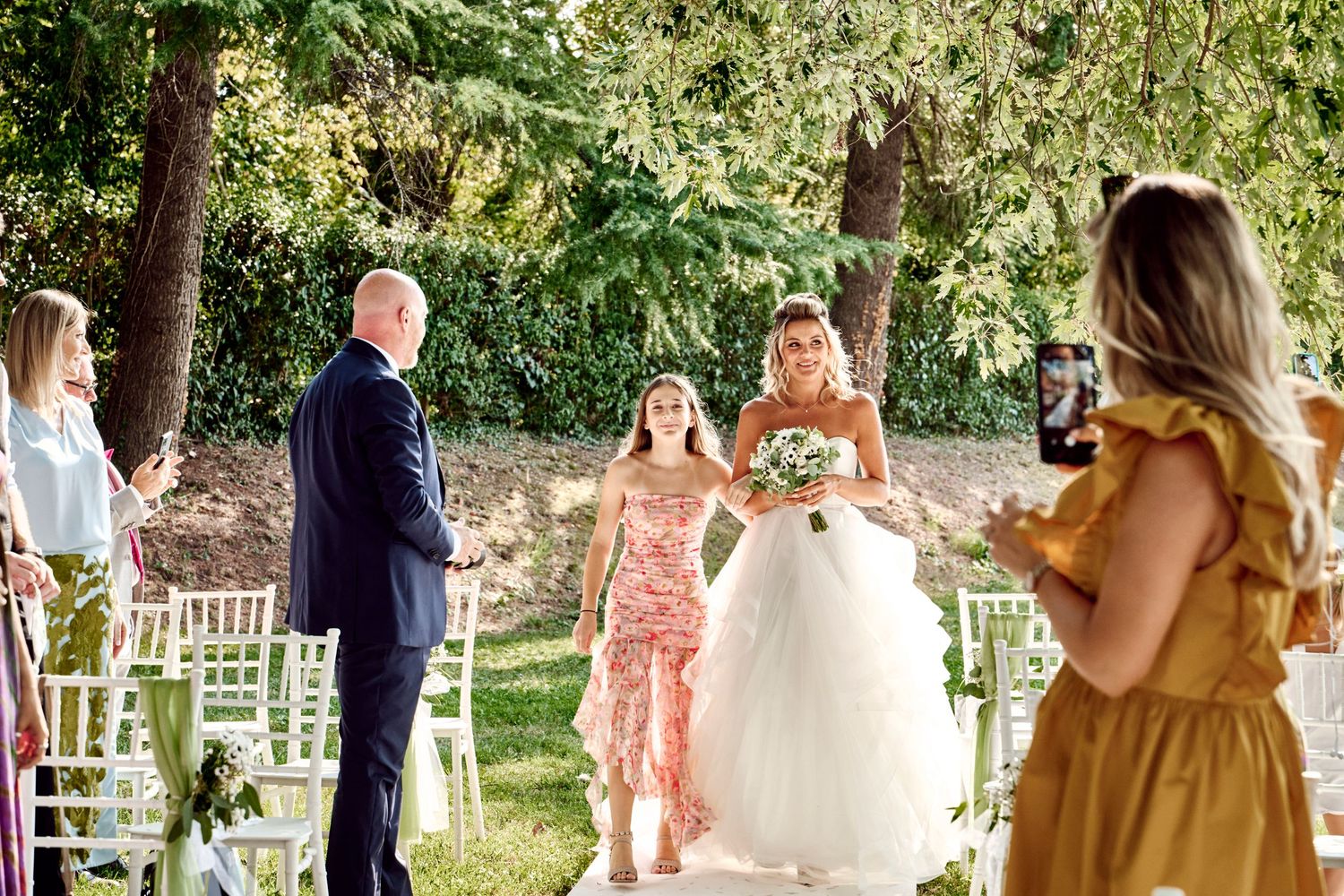 Bride walks down outdoor aisle with escort at garden wedding ceremony.