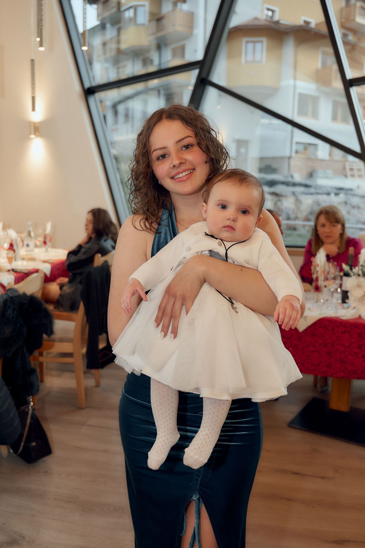 Smiling young woman in teal dress holding a baby in white dress at a baptism reception.