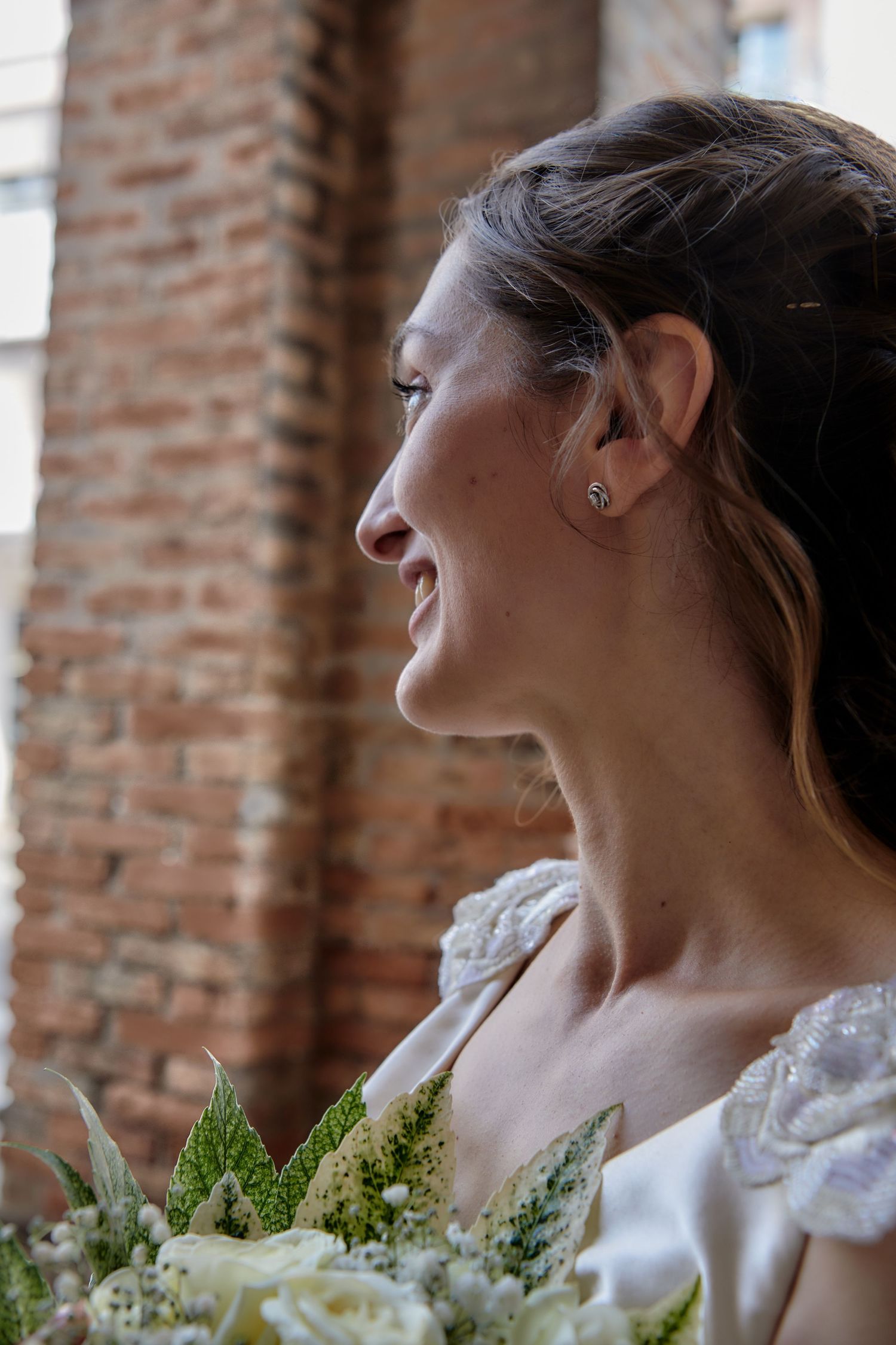 Smiling bride in profile holding white floral bouquet against a rustic brick wall background.