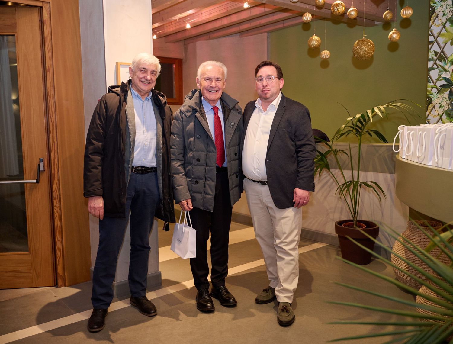Three older men posing together in a hotel lobby decorated with golden ornaments.