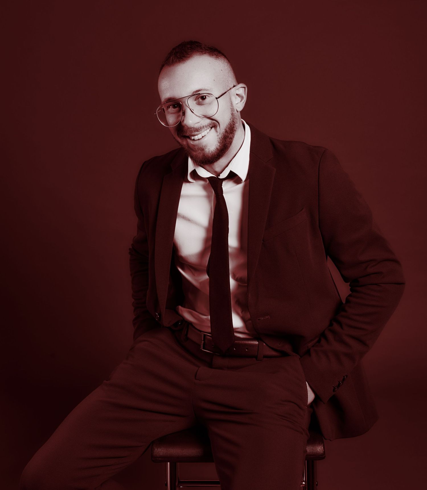 Smiling bearded man in suit and tie seated on stool against dark red background.