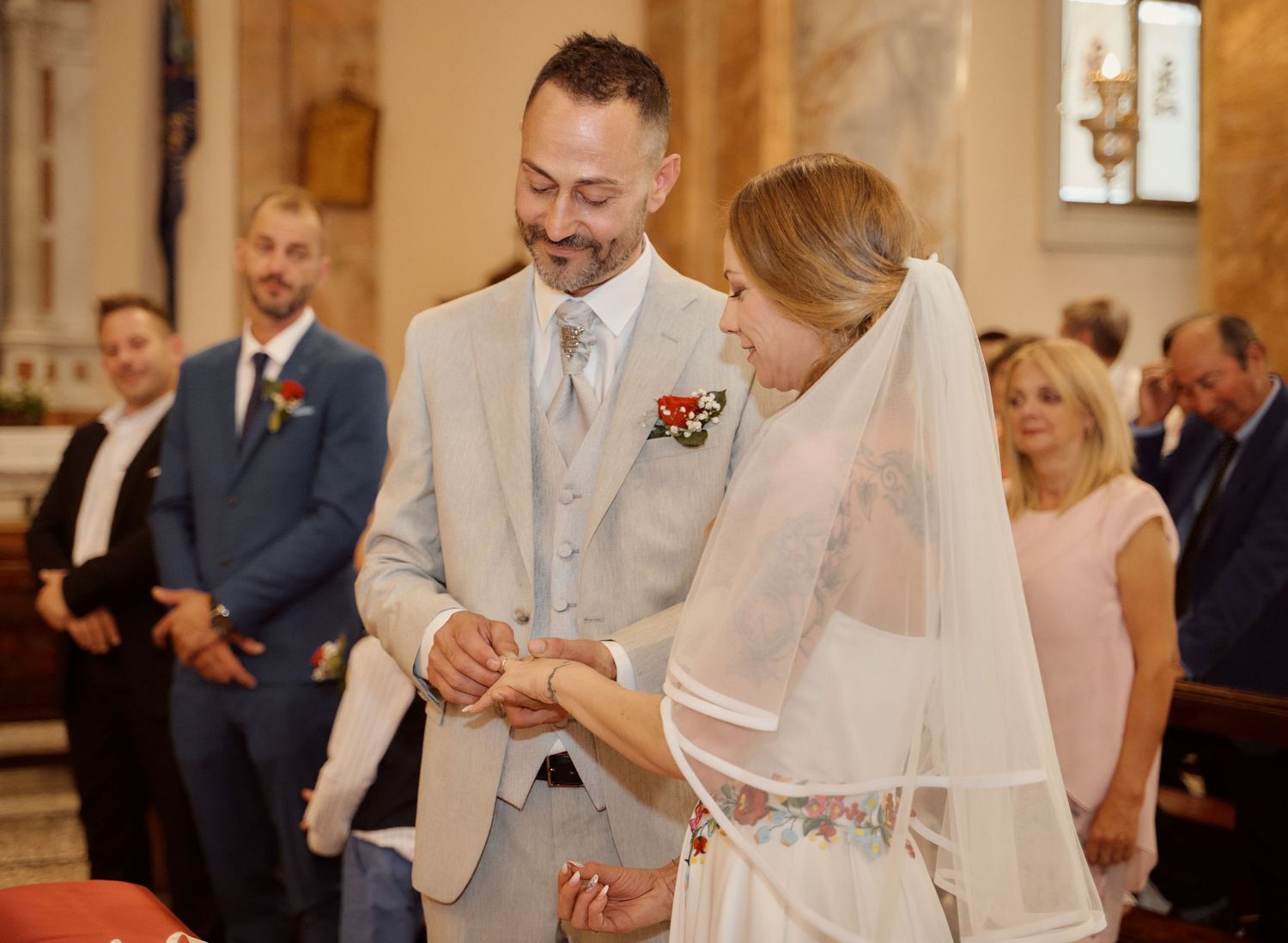 Groom smiles as bride places ring on his finger at church ceremony.
