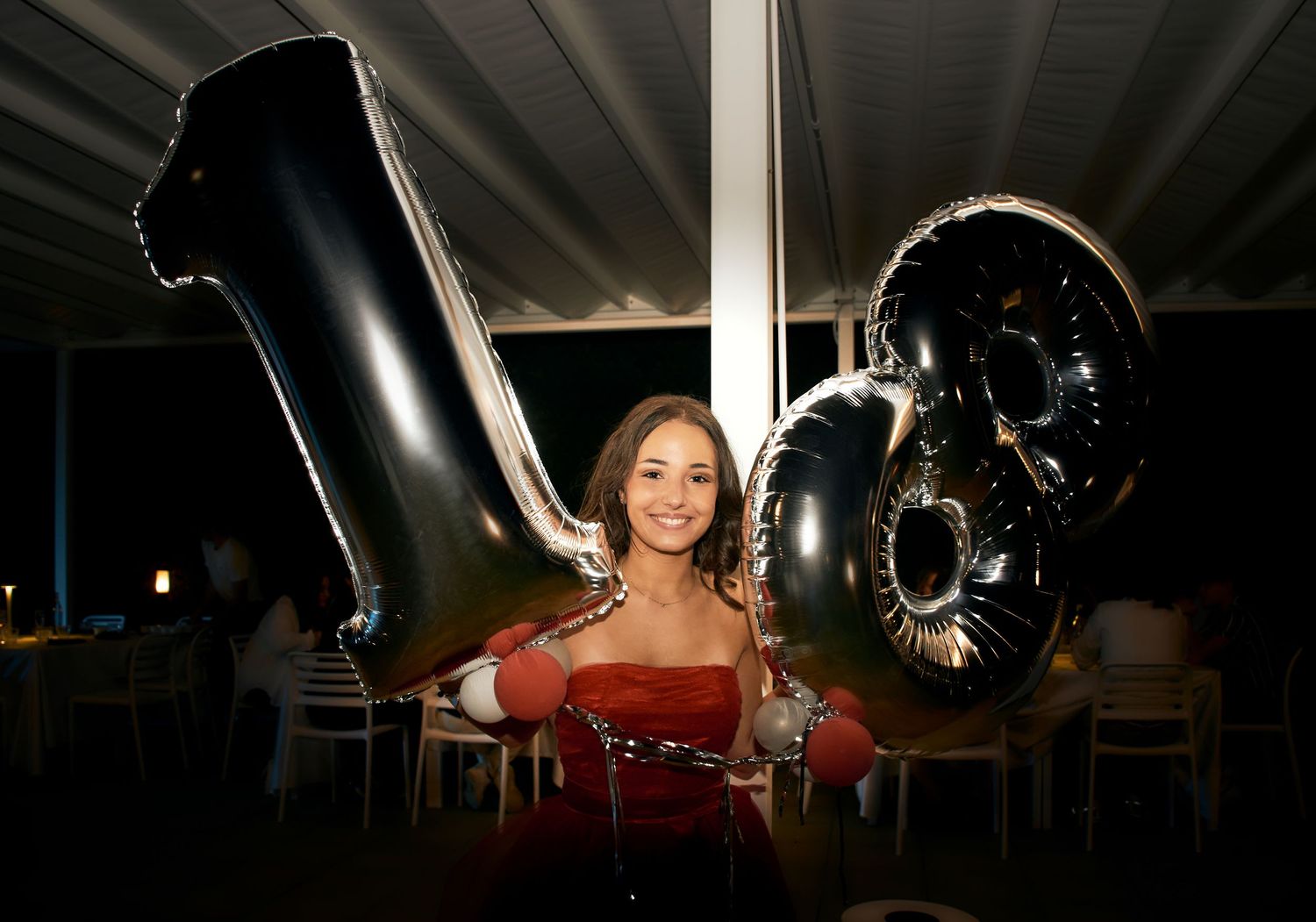 Young woman in red dress poses with large black number 18 balloons at a birthday party.