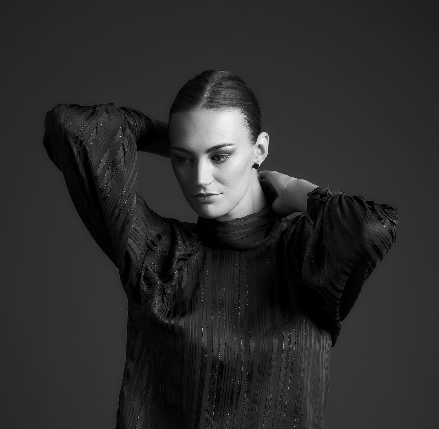 Elegant woman in black pleated dress poses gracefully against dark background in black and white fashion portrait.