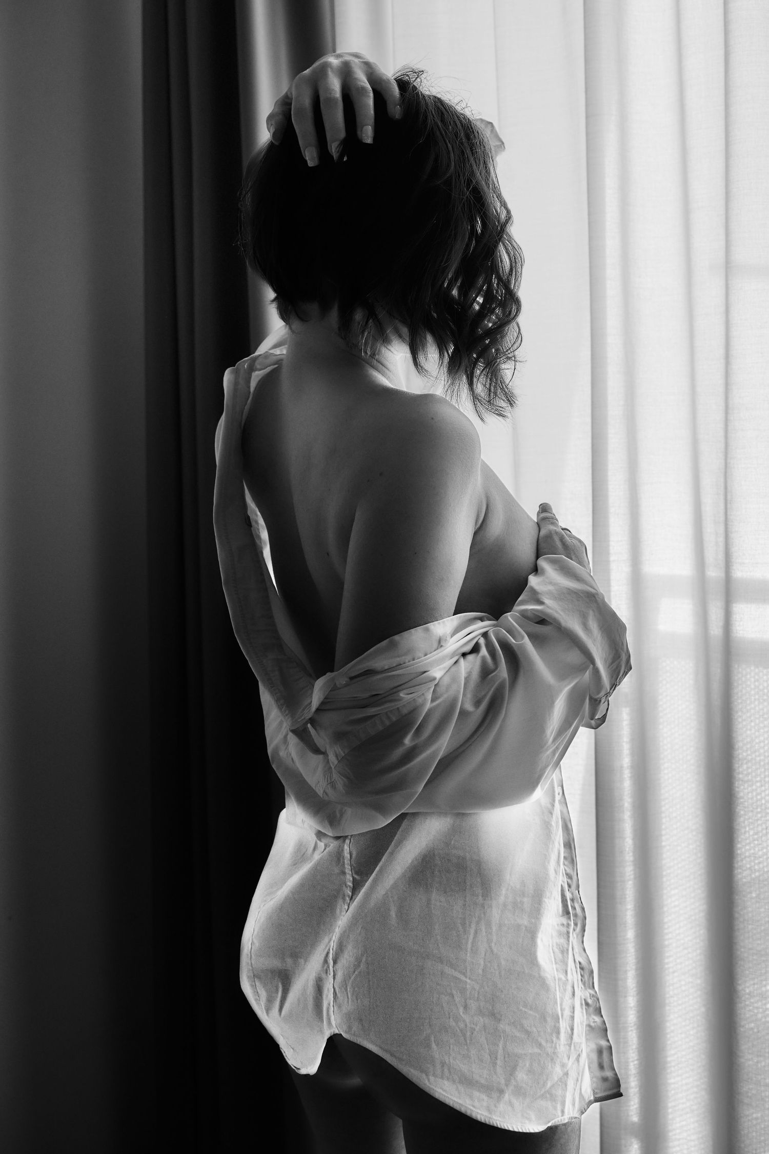 Black and white silhouette of woman in open white shirt looking out curtained window.