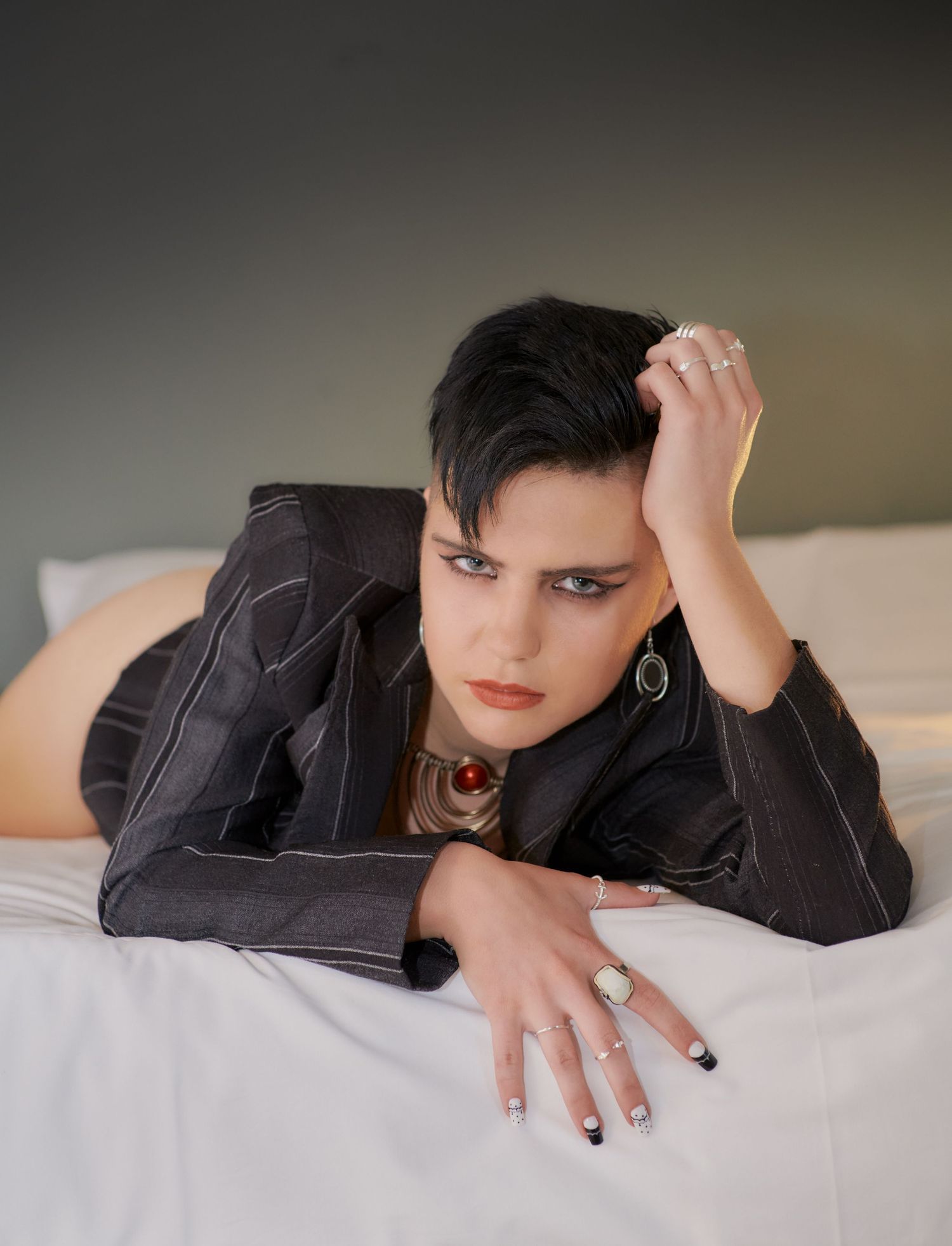 Short dark-haired woman in gray blazer lying on white bed with bold jewelry.