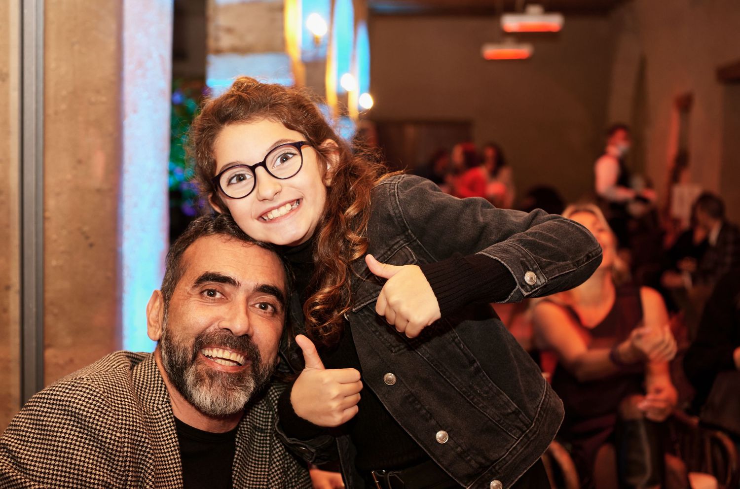 A bearded man smiles with a young girl wearing glasses giving a thumbs up at a lively event.