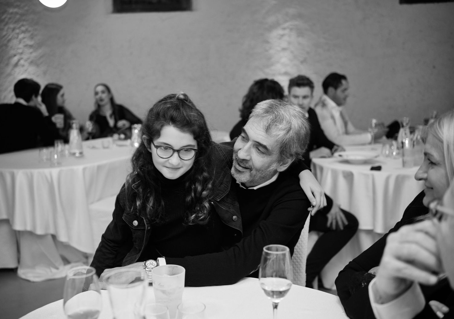 A black and white photo of a young girl with glasses and a man sharing a moment at a dinner table.