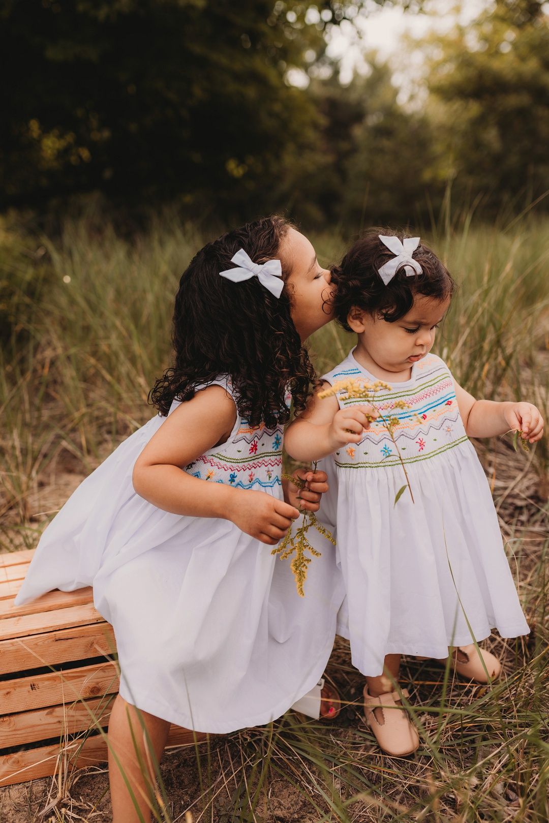 Two young sisters in matching white dresses share a sweet kiss outdoors in a field.