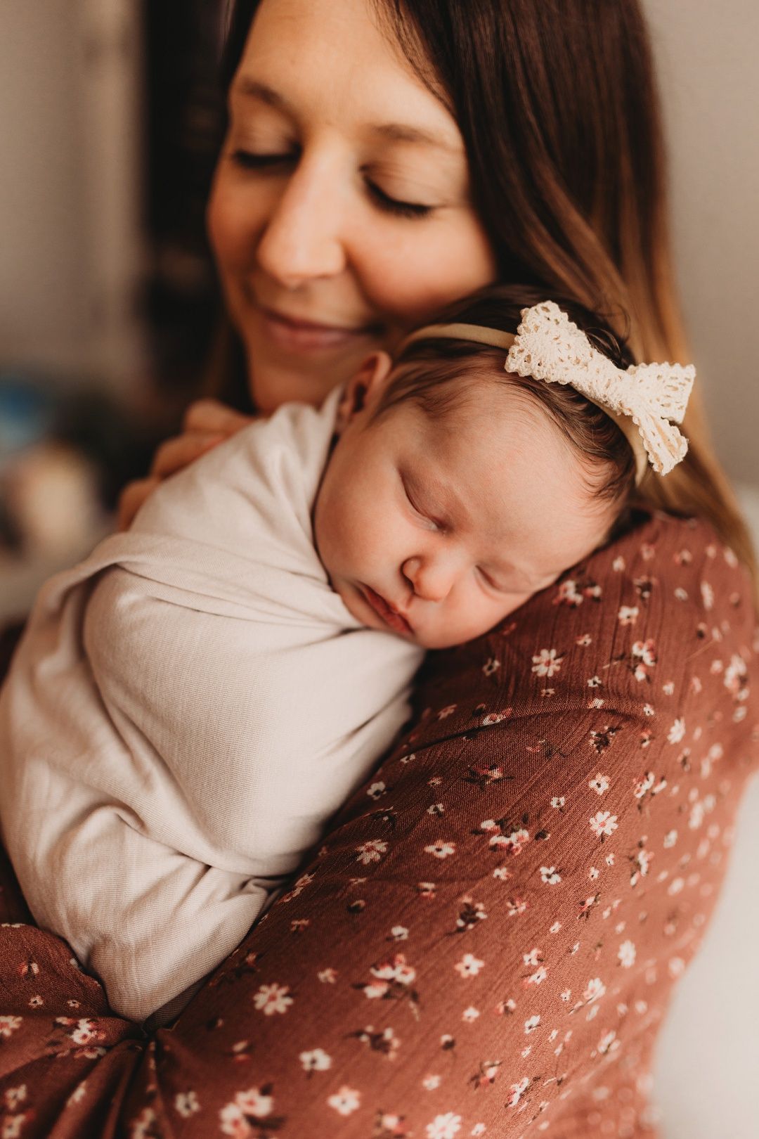 Dark-haired mother in floral dress smiles down at sleeping swaddled newborn with bow headband.