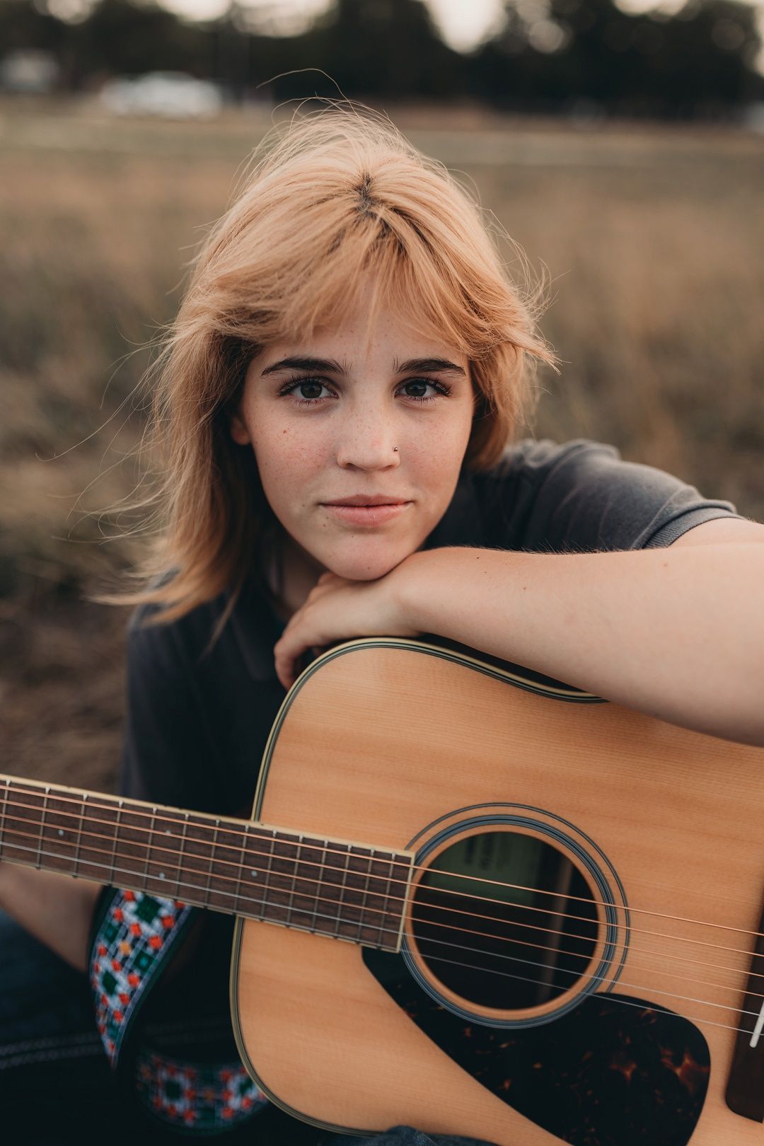 Blonde teen girl resting chin on acoustic guitar while sitting in golden field for senior photos.