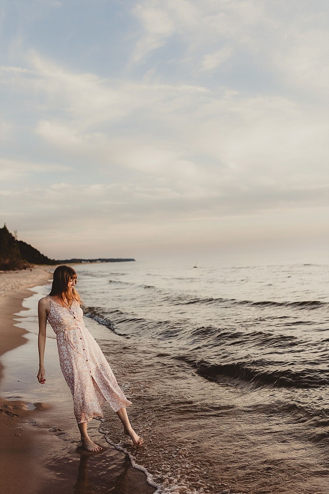 Young woman in floral sundress walking along shoreline at dusk, waves gently washing over her feet.
