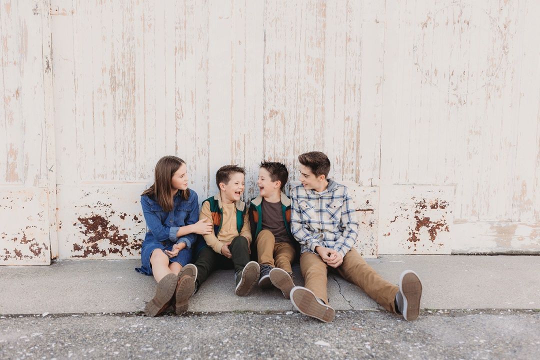 Four siblings sitting together laughing against a rustic white wooden wall outdoors.