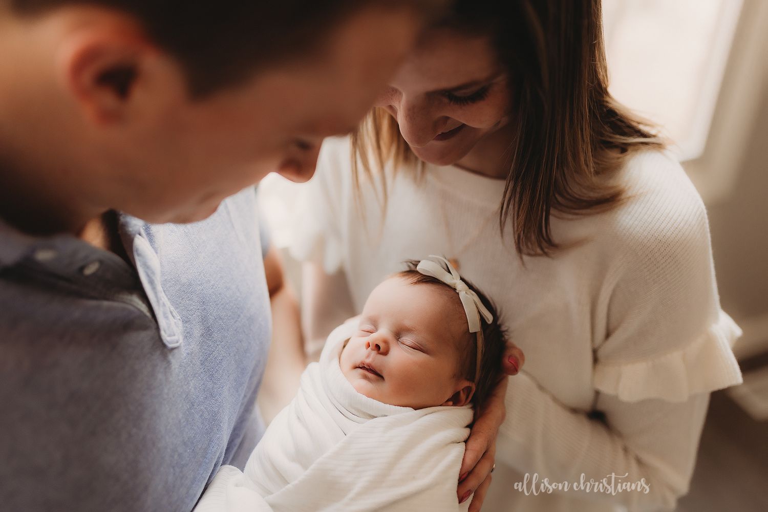 New parents gazing lovingly at sleeping swaddled newborn baby near window at home.