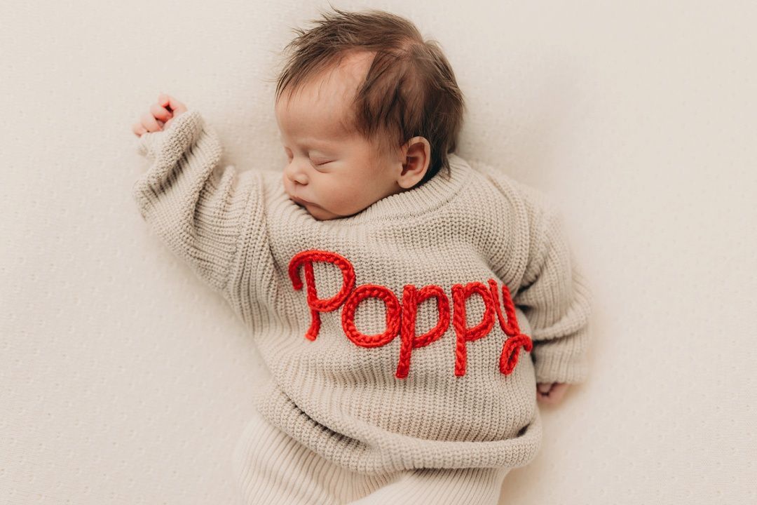 Sleeping newborn baby wearing a personalized beige knit sweater with red embroidered name Poppy.