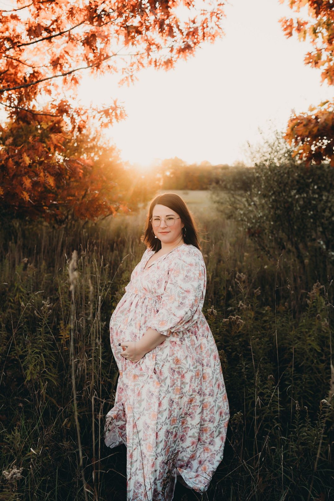 Pregnant woman in floral dress stands in golden sunset light among tall grass.