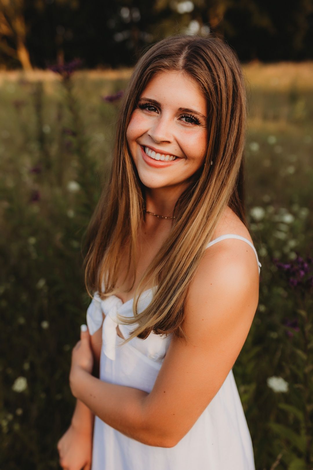 Smiling teenage girl in white dress standing in a wildflower meadow during golden hour.