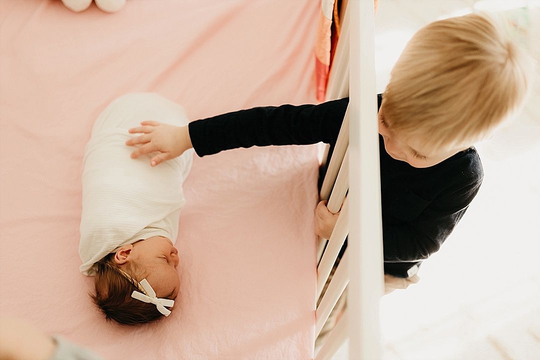 A young toddler reaches through crib slats to gently touch a sleeping newborn baby girl.
