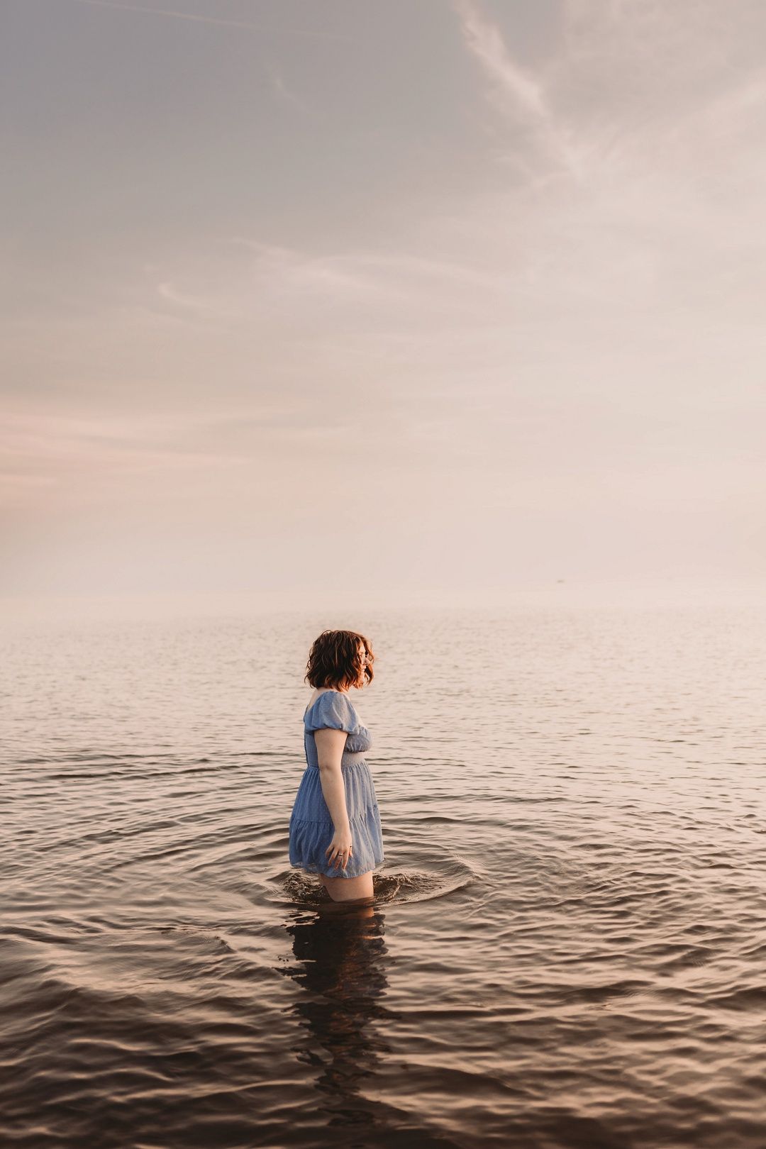 Woman in blue dress standing in calm ocean water at sunset.