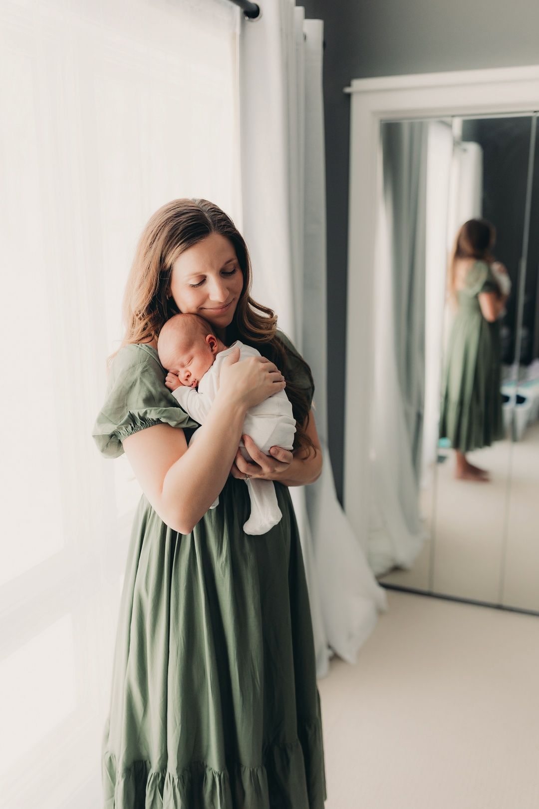 New mother in green dress holding sleeping newborn baby near bright window with mirror reflection.