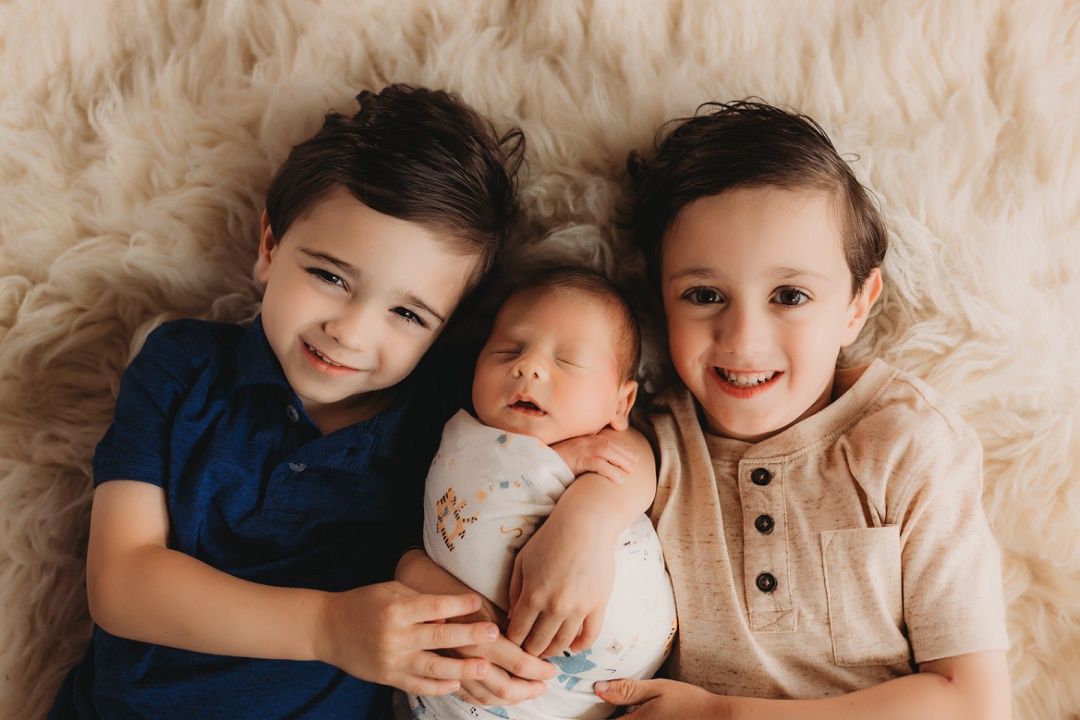 Two smiling boys pose with their newborn sibling on a fluffy cream blanket.