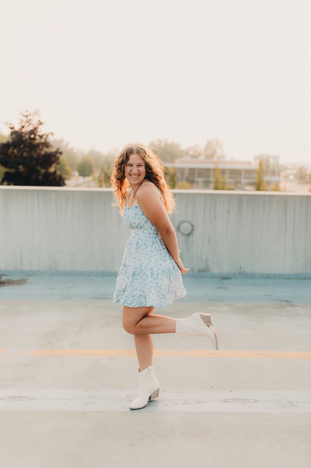 Young woman in blue floral dress and white boots poses playfully on rooftop.