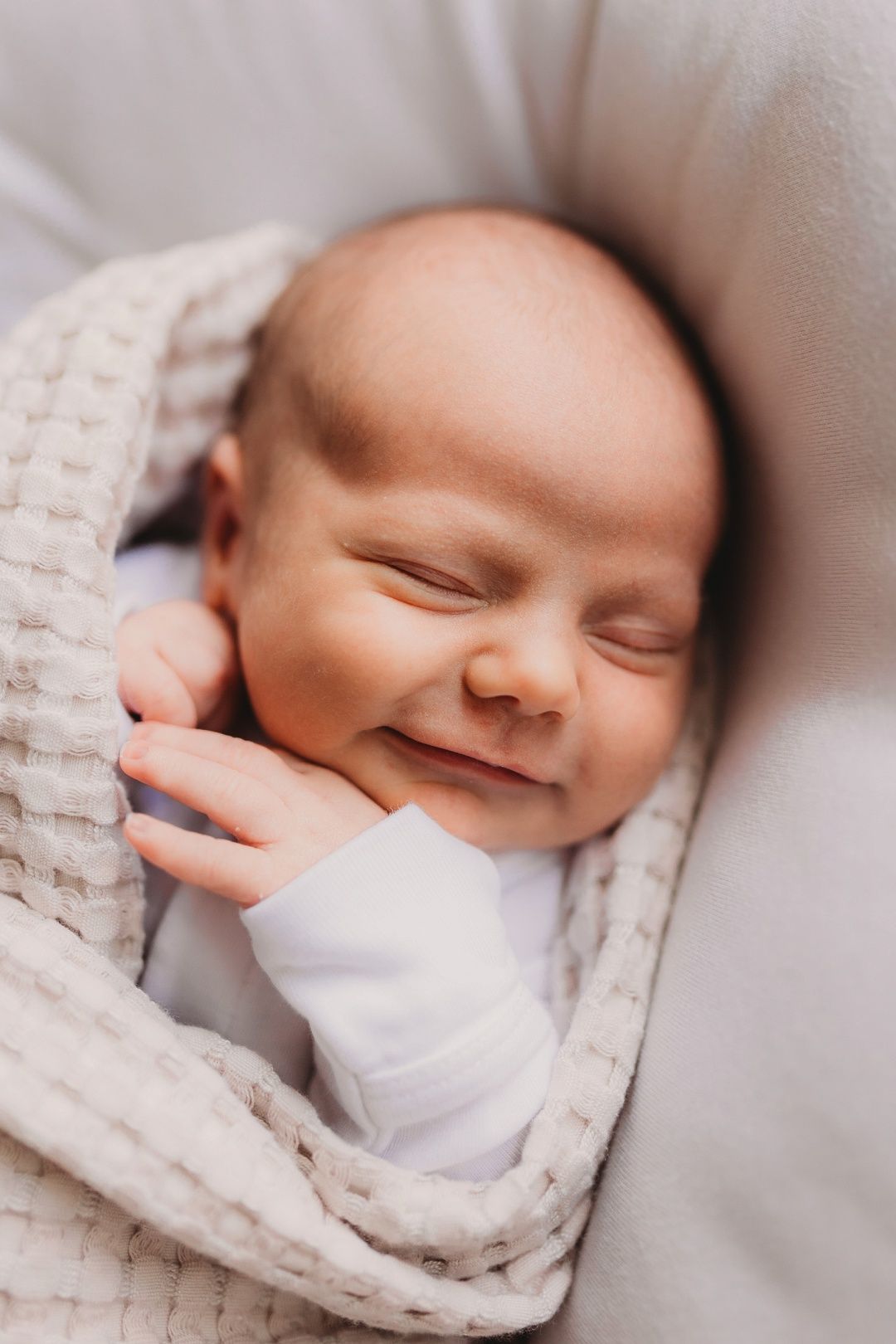 A peaceful newborn baby smiles softly while wrapped in a cozy cream blanket.
