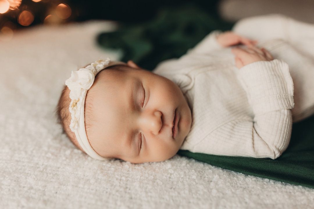 Sleeping newborn baby girl in white outfit wearing floral headband on soft white blanket.