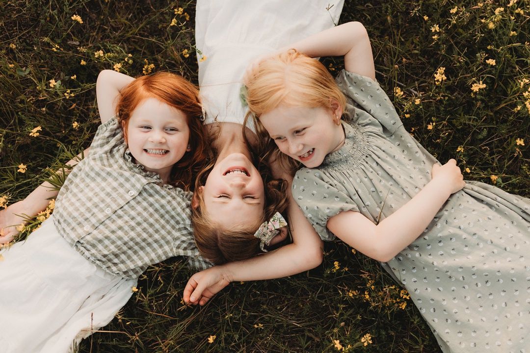 Three red and blonde-haired sisters lying in grass laughing together outdoors.