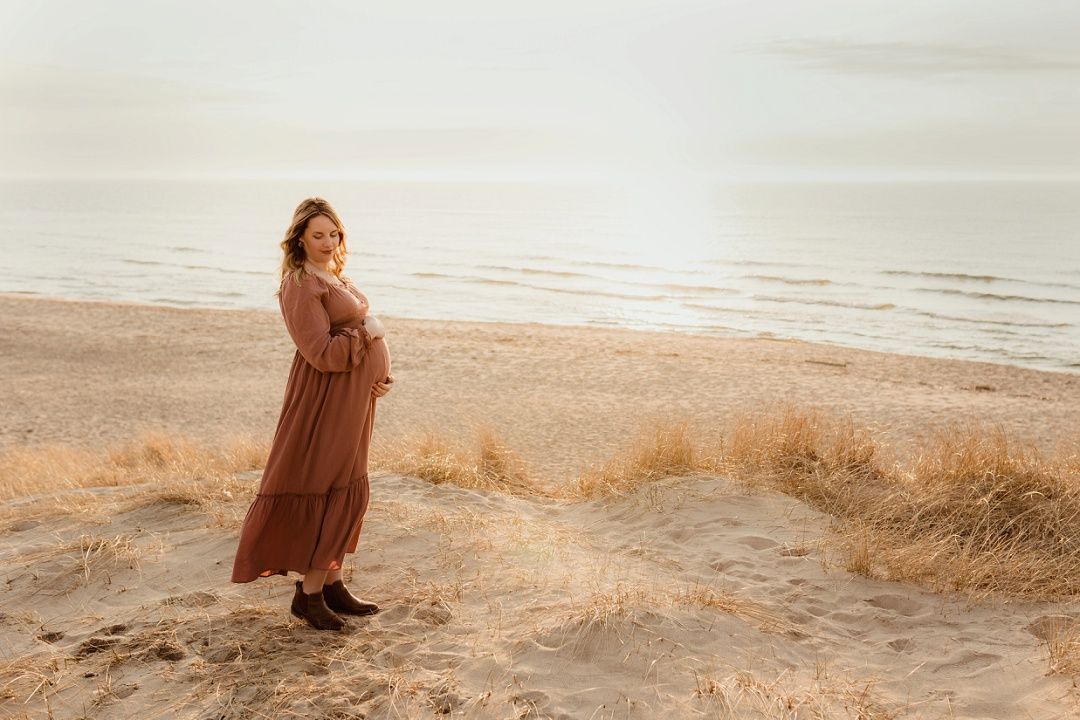 Pregnant woman in brown maxi dress standing on sandy beach at soft overcast light.