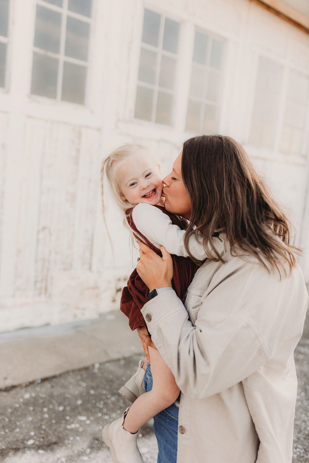 Mother kissing her laughing toddler daughter outdoors near a white vintage building.