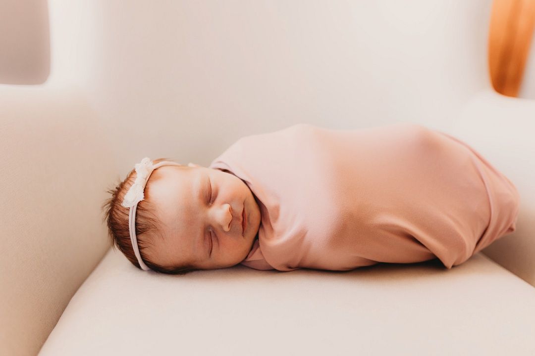 Swaddled newborn baby in pink wrap with headband sleeping peacefully on white surface.