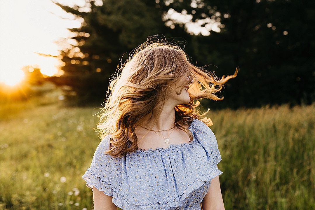 Young woman with flowing blonde hair in blue off-shoulder dress twirling in sunlit meadow at sunset.