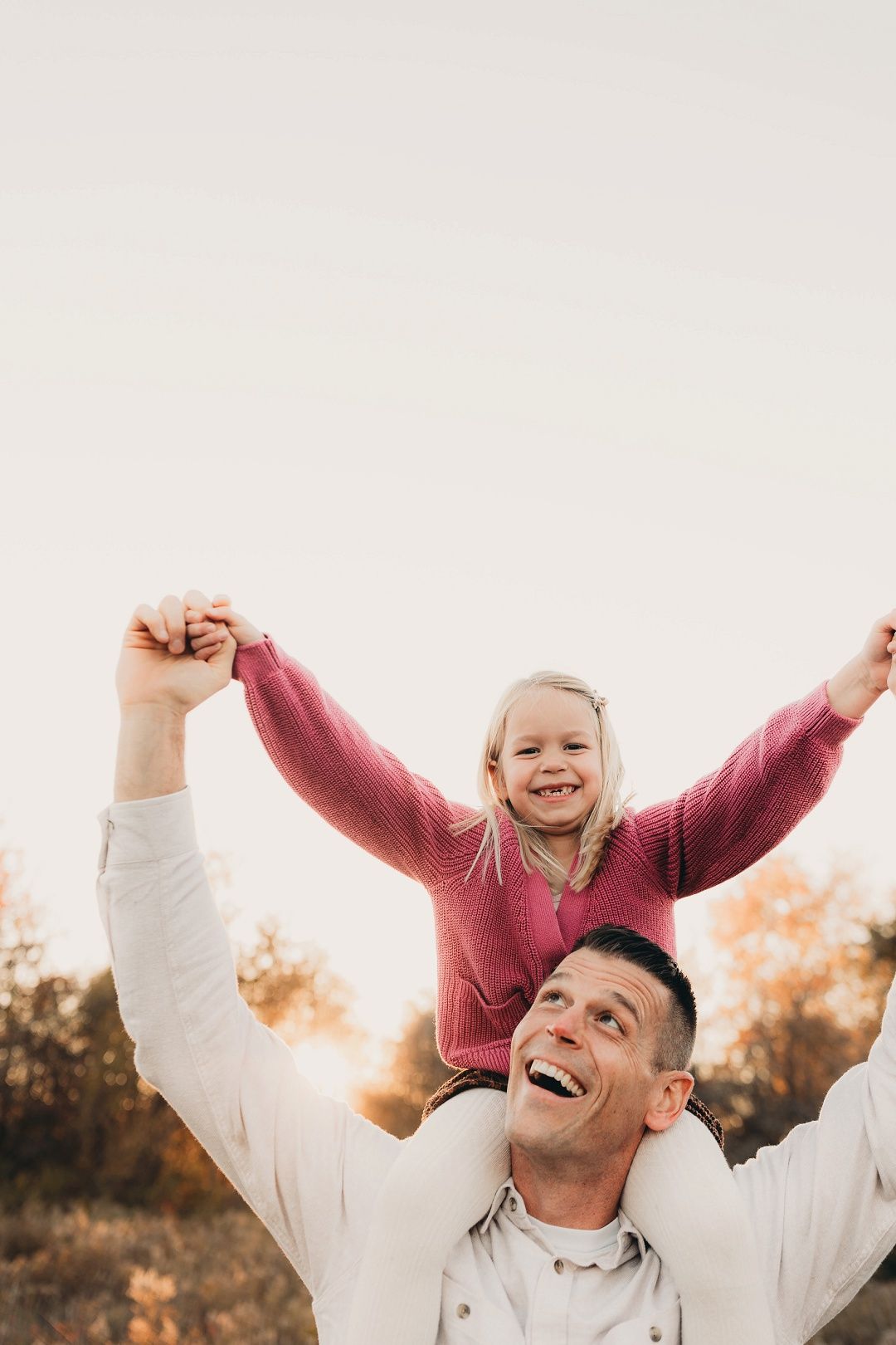A laughing girl spreads her arms wide while sitting on her fathers shoulders outdoors.