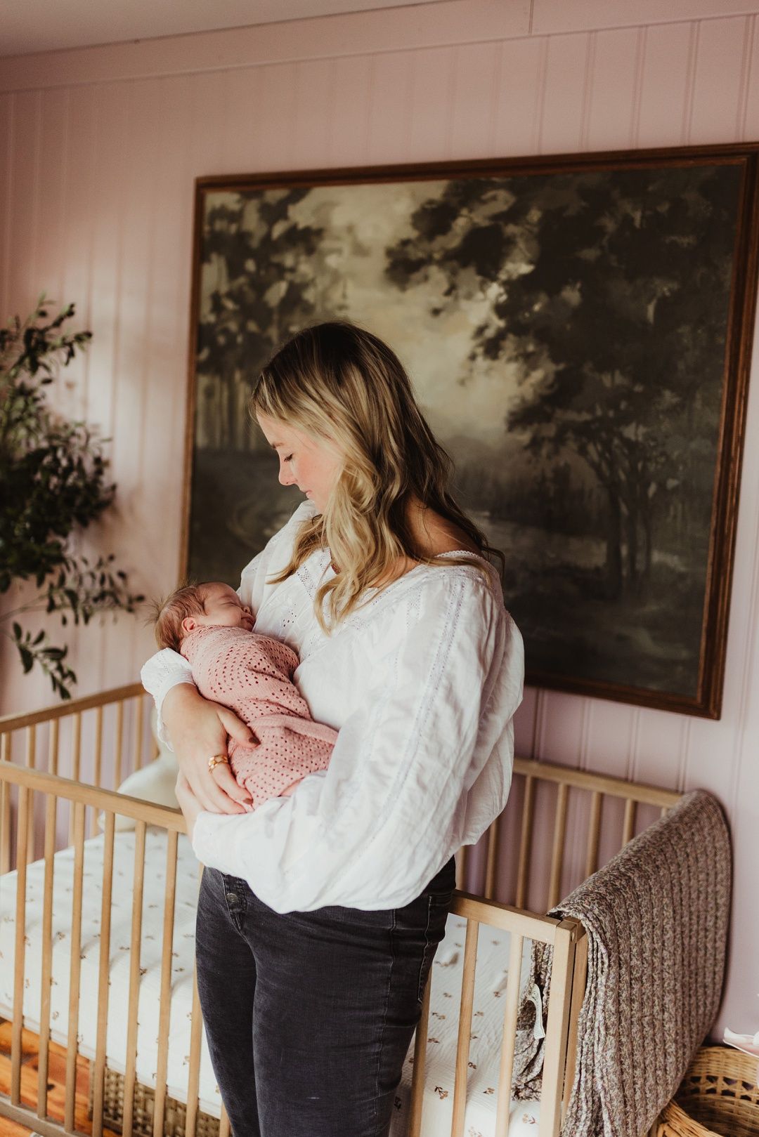 Blonde mother holds pink knit-wrapped newborn in a pink nursery with vintage artwork.