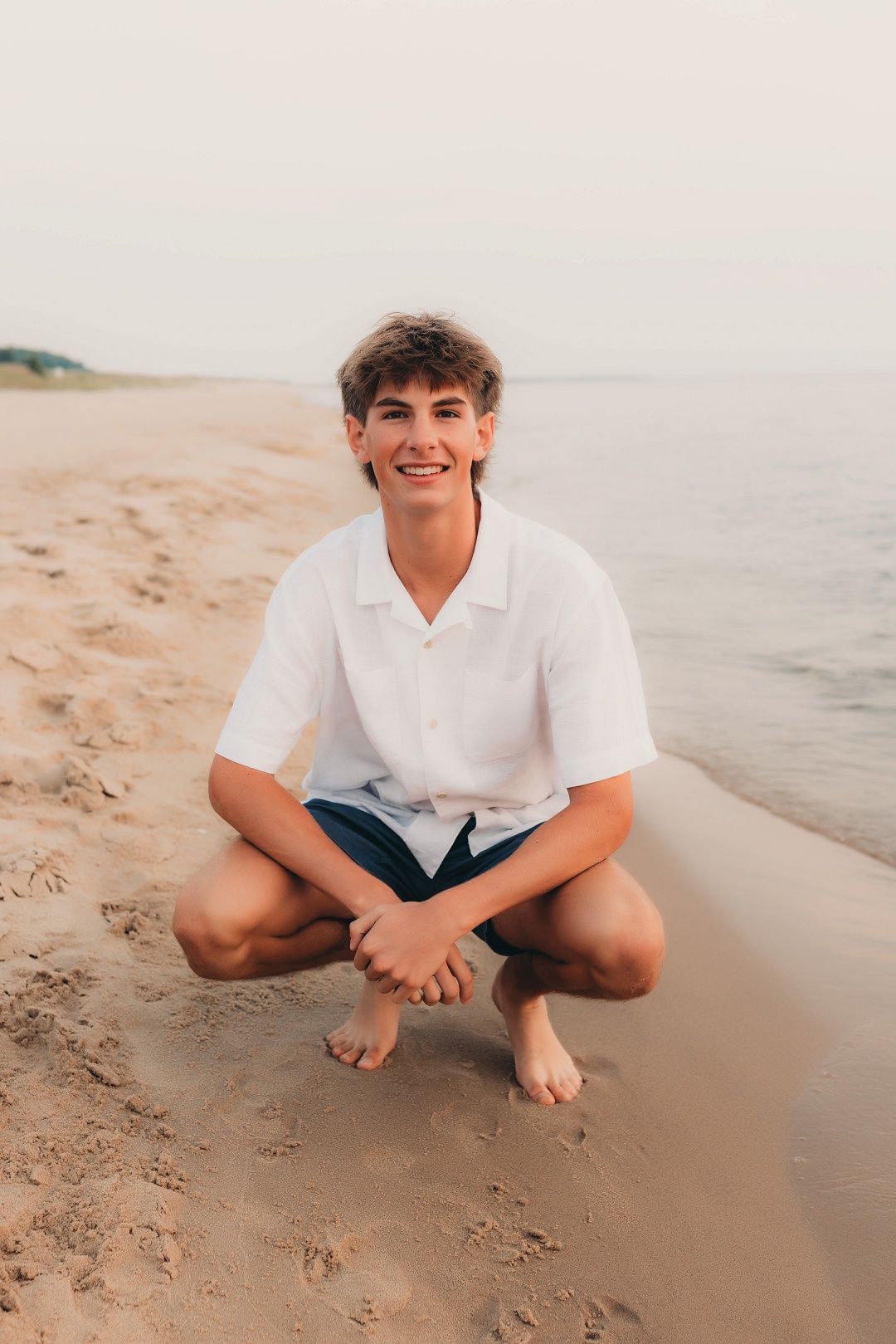 Teen boy in white polo shirt crouching barefoot on sandy beach shore smiling for senior photos.