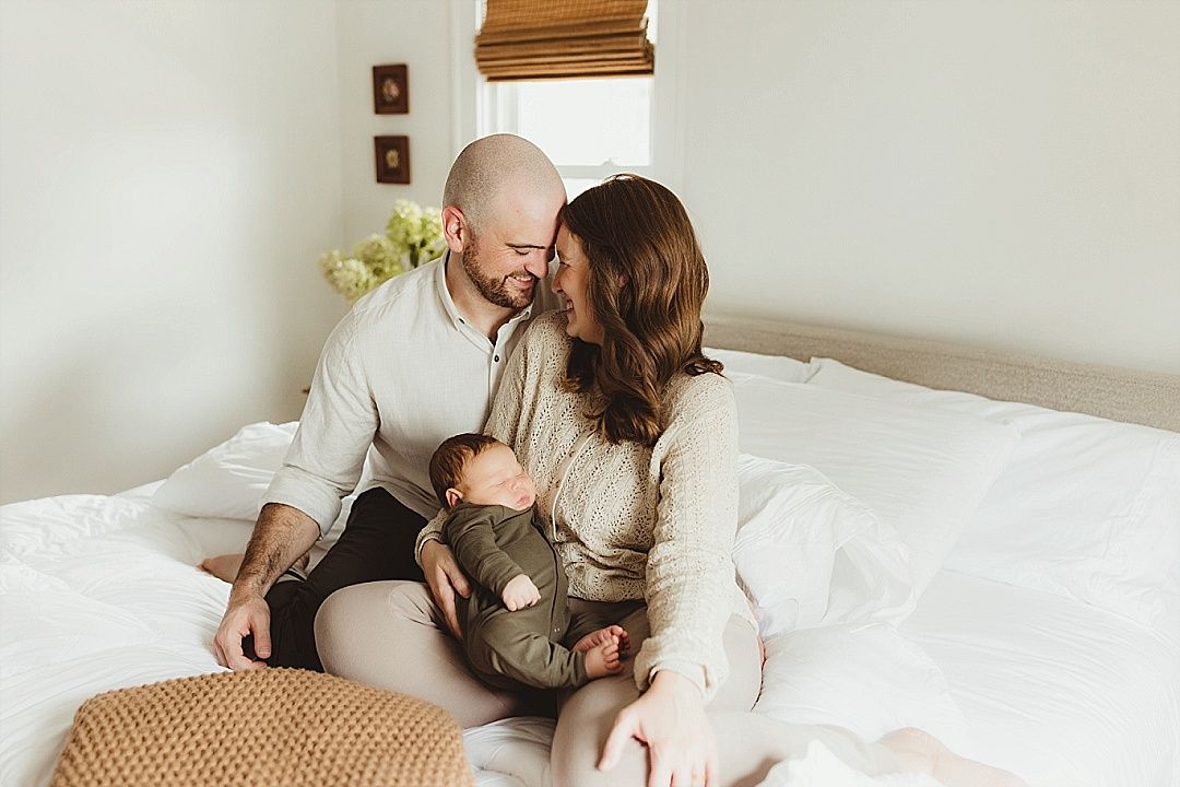 Happy couple sits on white bed smiling at each other while holding their newborn baby.