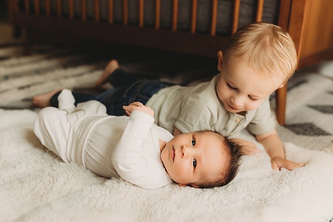 A smiling toddler cuddles next to their newborn sibling lying on a soft nursery rug.