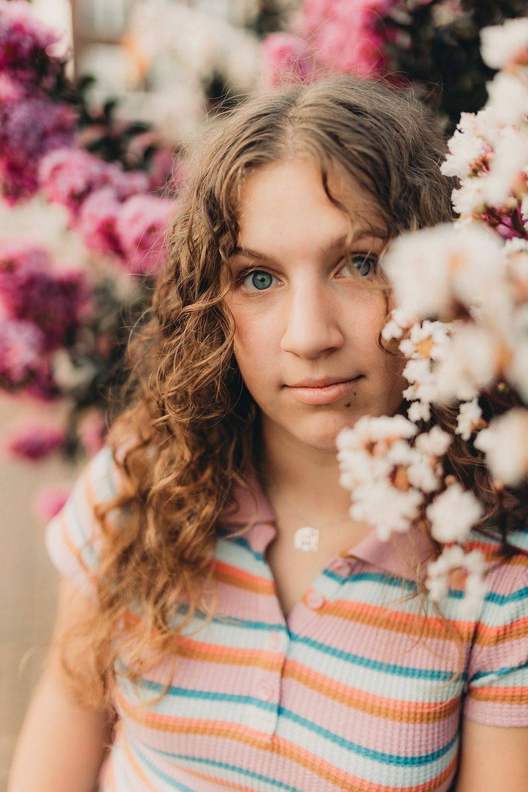 Young woman with curly hair posing near pink and white flowering bushes.