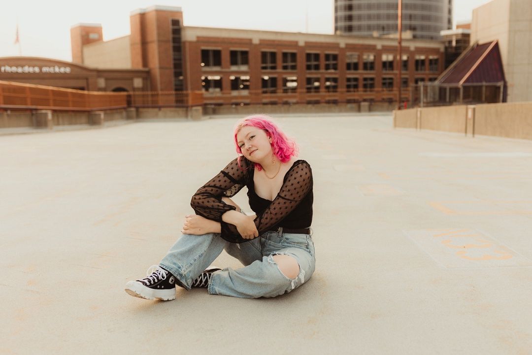 Young woman with pink hair sitting on urban rooftop in ripped jeans and black lace top.