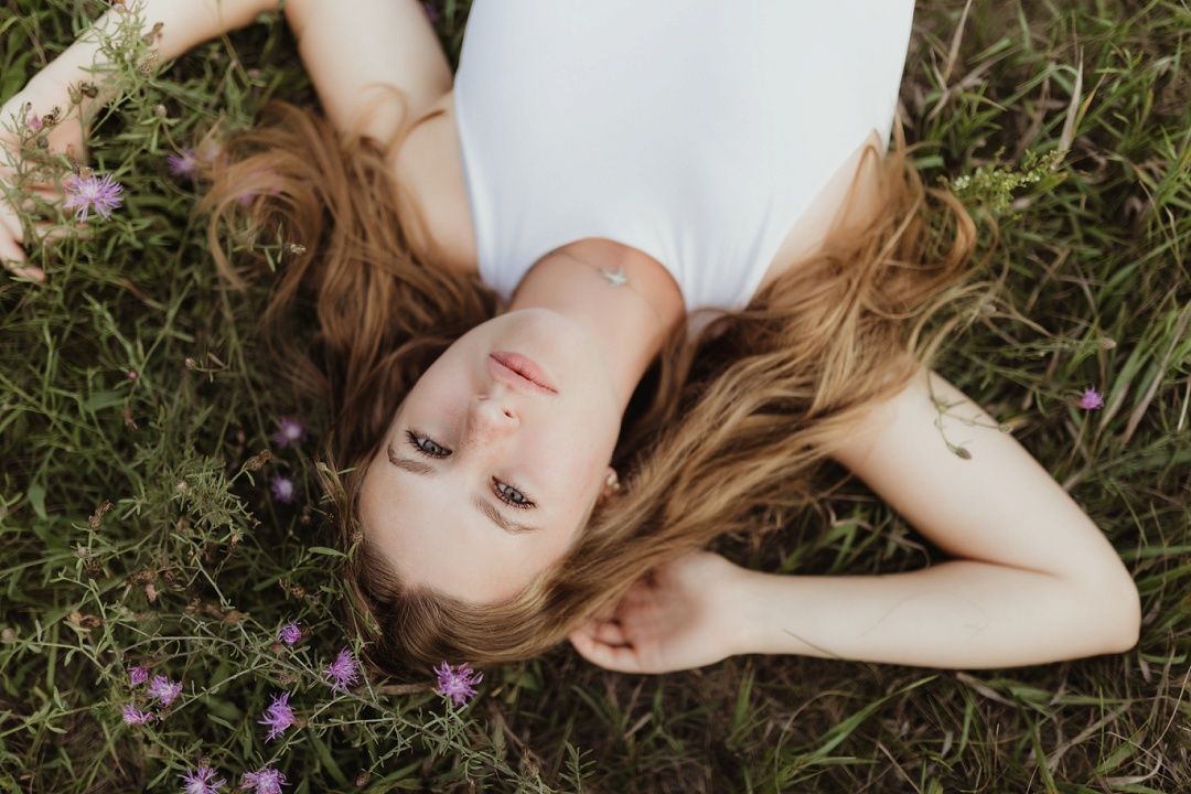 Young woman with long hair lying in wildflower meadow, eyes open, relaxed outdoor portrait.