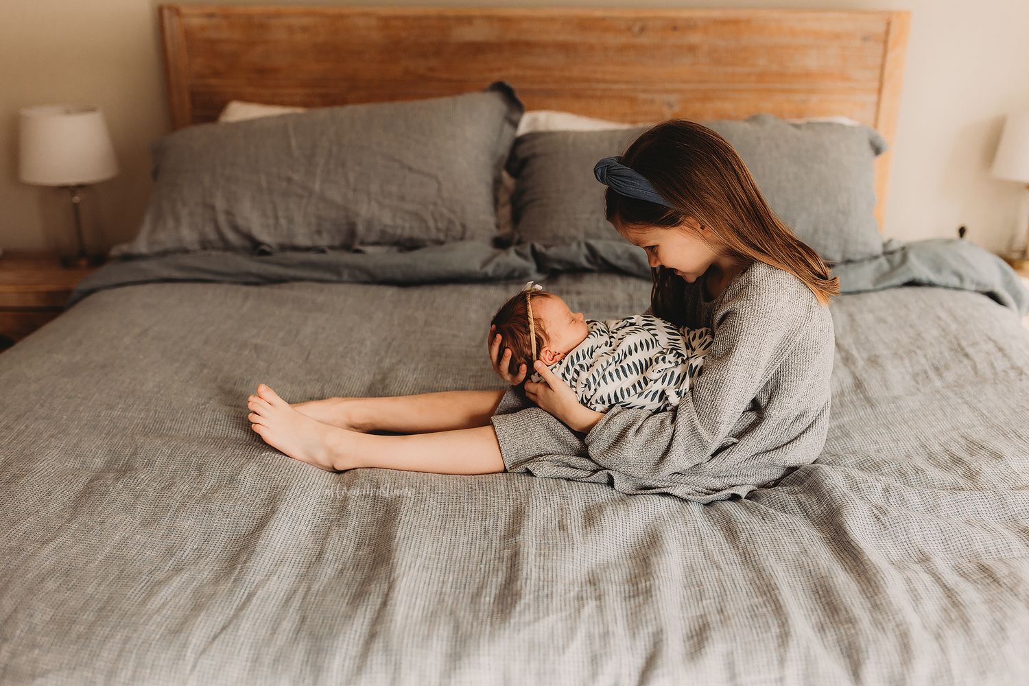 A mother sits on a gray linen bed lovingly holding her newborn baby close to her chest.